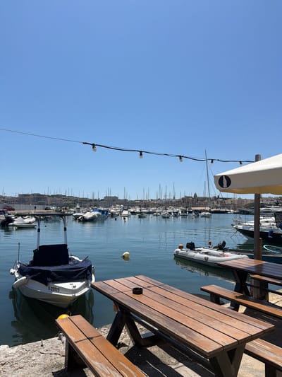 The image shows a sunny day at the Sliema Ferries in Gżira, Malta. In the foreground, two wooden picnic tables with benches are set up on a paved area. One table has an ashtray on it. To the left of the tables, a small white boat with a navy blue cover is docked. The boat has "S-25925" written on its stern. In the background, a harbor is filled with numerous sailboats and other vessels. Beyond the harbor, the city skyline of Sliema and Valletta is visible, characterized by historical buildings and architecture. The sky is clear and blue. An umbrella with a black circle and white shape logo is visible to the right of the tables. A string of lights hangs across the top of the frame.