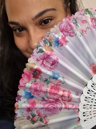 In Orlintsi, Bulgaria, a woman is photographed indoors, partially obscuring her face with a floral fan. Her dark hair frames her face, and only her eyes and forehead are clearly visible. She has dark brown eyes and well-defined eyebrows. The fan, adorned with pink and blue flowers, is held open and positioned to the right of her face, covering her nose and mouth. The fan has a white, decorative edge. The background is indistinct, suggesting an indoor setting.