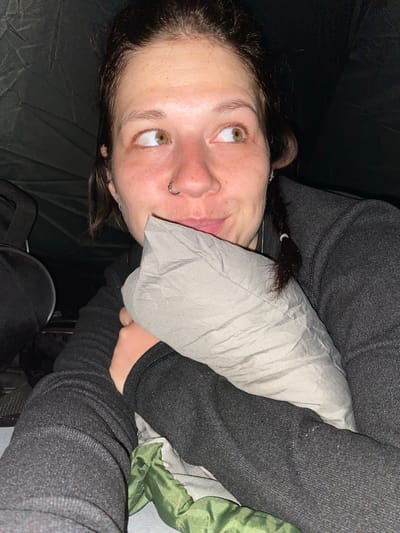 The image shows a close-up, low-angle shot of a young Caucasian woman lying down, holding a gray pillow. She has light skin with freckles and is looking to her right. Her hair is dark brown and pulled back in a braid on the right side, with some wisps framing her face. She is wearing a dark gray hooded jacket. A nose ring is visible on her septum. The background is dark and appears to be the interior of a tent, with a dark green sleeping bag or fabric visible at the bottom. A black bag is partially visible to the left of the frame. The lighting is dim, suggesting it is nighttime or a dark indoor environment. The scene is likely in Абзаново, Russia, given the context.