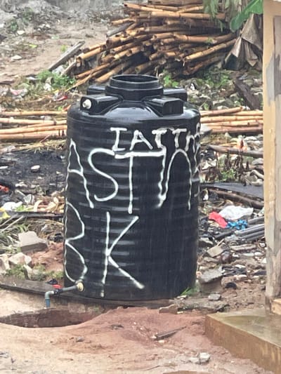 A black plastic water storage tank rests on the ground amidst debris in an outdoor setting, likely in Abraka, Nigeria. White spray paint decorates the tank with the lettering "LAT SIK". The surrounding environment features bamboo poles, discarded materials, and dirt. The tank is positioned near a weathered concrete or earthen wall structure. A small blue and red valve is connected to the tank.