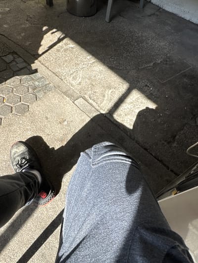 The image shows a person's legs and feet, along with a shadow cast by an unseen object, likely a window frame or building structure. The scene appears to be outdoors, on a paved surface such as a sidewalk or patio, with patches of sunlight and shadow indicating a bright day.

The subjects are a person's lower body, visible from the thighs down to the shoes. The person is wearing dark-colored pants and dark athletic shoes with red accents. One shoe is in sharp focus, revealing a brand name with red lettering, though the full name is not clearly legible. The other leg and foot are partially visible, also in shadow.

The setting is a textured, outdoor surface. The ground is a mix of concrete and what appears to be hexagonal pavers. Sunlight is casting a distinct rectangular shadow with crossbars across the ground, suggesting the light source is coming through a window or similar opening. A small portion of a white wall or building is visible in the upper right corner, and a dark, cylindrical object is present on the left edge of the frame, possibly a trash can or planter.

The strong contrast between light and shadow suggests it is daytime, likely mid-morning or afternoon, given the angle of the sun. The bright sunlight and clear shadows indicate fair weather. There is no explicit indication of an activity or event taking place, other than the person is likely standing or sitting outdoors. No discernible emotions are conveyed. The location context of Sankt Wendel, Germany, provides a geographical anchor but doesn't offer specific visual cues within the image itself.