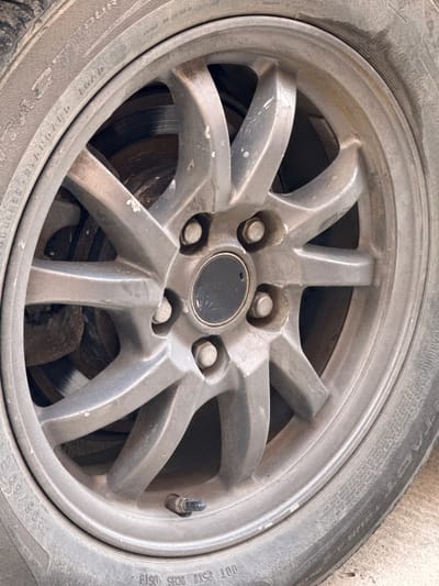 The image shows a close-up of a car wheel, specifically the rim and tire. The wheel is silver and has a five-spoke design. The tire is black and appears to be slightly worn, with some dirt and scuff marks visible. The wheel is mounted on a car, and the brake rotor is visible through the spokes of the rim. The background is out of focus, but it appears to be a concrete surface.
There is text visible on the tire, including "RELATION PRESSURE 350 KPA", "WARNING", "MAX LOAD 630 KG (1389 LB)", and "DOT P5X1T OCHS 0519 205/60R15". These markings provide information about the tire's specifications, including its maximum inflation pressure, load capacity, and manufacturing date.
The image appears to be taken outdoors, likely in a garage or parking area, given the concrete surface and the presence of a car. The lighting suggests it could be daytime, but no specific time of day can be determined. There are no people or animals visible in the image. The overall impression is one of a functional, but not new, car part. The wear and dirt suggest the car is in regular use. The location context provided is Mastaga, Azerbaijan.