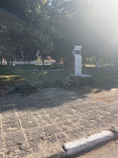 The image captures an outdoor scene in the city of Gradina, Bulgaria, likely during the daytime given the bright sunlight. In the foreground, a paved pathway made of square, grey stone tiles is visible, with a white concrete curb running along its edge. The pathway is partially shaded by the overhead sun.
Further back, a patch of green grass forms a lawn. Along the edge of the lawn, a low decorative border of white, pointed stones is present. In the middle of the lawn, a white, abstract-shaped monument or sculpture stands on a square concrete base. The sculpture has a darker rectangular plaque on its front.
Around the base of the sculpture and along the lawn's edge, a cluster of flowering plants with small purple and pink blossoms is growing. Beyond the lawn, a line of trees with dense green foliage forms a backdrop. The sunlight is strong, creating lens flare and casting bright highlights in the upper right portion of the image. In the background, beyond the trees, hints of buildings are visible. There are no people present in the frame.