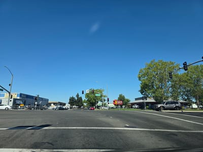 This image captures a wide-angle view of a street intersection on a clear, sunny day. The sky is a vibrant, cloudless blue, suggesting a warm, pleasant atmosphere. The foreground is dominated by asphalt, with white lane markings indicating a multi-lane intersection.
On the left side of the road, a commercial building is visible with signage that includes "E-Z SMOOG" and "IN & OU VE ALSO LA HYBRIDS". Next to it, another building displays "ANSMISSIONS Auto Tecy Repor" and "WE ALSO DO 'STAR' SWOGS". Several cars are parked and driving in this area.
In the center of the intersection, traffic lights are visible, with one showing a red light. Street signs indicate "Martin Blvd". A red car is stopped at the intersection.
On the right side of the intersection, a prominent "Carls Jr." restaurant sign with its distinctive star logo is clearly visible. Another building, possibly associated with the restaurant, is also present. A grey SUV is parked in front of it, and several other vehicles are in the background. Large, leafy trees provide shade and add greenery to the scene. The overall setting is a typical suburban commercial area in the United States, likely during daylight hours due to the bright sunshine and strong shadows. There are no people clearly visible in the image.