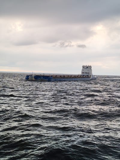 A cargo barge moves across choppy water under an overcast sky. The barge is long and flat with a raised superstructure at the stern, containing windows and what appears to be a navigation bridge. The water is a deep, dark blue with numerous waves, suggesting a windy or rough condition. The sky is filled with gray clouds, with some patches of brighter light indicating the sun may be breaking through or is low on the horizon. There are a few small, dark shapes in the sky that could be birds. The barge has some visible text on its side. The scene suggests a utilitarian setting, likely a body of water used for shipping or transport. The weather appears cool and possibly blustery, giving the overall scene a somewhat dramatic or somber mood. The time of day is unclear due to the overcast conditions, but the light suggests it could be morning or late afternoon.