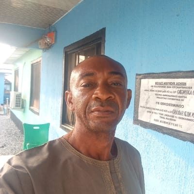 An adult male with dark skin and a bald head stands outdoors in Koko, Nigeria, directly facing the camera with a neutral expression. He is wearing a brown long-sleeved traditional garment featuring embroidered trim around the neckline.
The man is positioned in the foreground, with a building's bright blue wall forming the immediate background to his right and behind him. A white rectangular plaque, mirrored and upside down in the image, is affixed to this blue wall. The legible text on the plaque indicates "MEDICAL RECEPTION STATION RECONSTRUCTED AND REMODELED" "FOR THE GLORY OF GOD AND THE GENERAL WELFARE OF KOKO NAVAL BASE," and states it was "COMMISSIONED BY: BRIGADIER GEN MD SARASO, ACTING COMMANDING OFFICER, 23 AMPHIBIOUS BATTALION, NIGERIAN ARMY" on "18 SEPTEMBER 2023." A dark-framed window is visible on the blue wall behind the man's head, to his left.
Further to the left, a second building with a lighter blue-green facade is visible, featuring an external air conditioning unit mounted on its wall. A green plastic chair rests on a gravel or stone-covered ground surface in front of this building. A grey corrugated roof overhang extends above the blue wall and the man, with an orange electrical box or meter suspended from it. The scene appears to be under natural daylight.