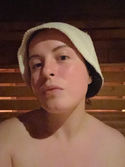 A person wearing a white sauna hat is shown in a close-up, slightly tilted view. The skin tone appears flushed or reddish. The background features vertical wooden slats, suggestive of a sauna interior. Some shadows are present, adding depth to the image. The focus is mainly on the face and upper body of the person.