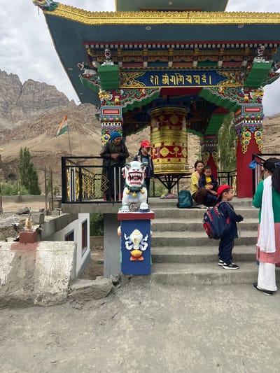 The image shows a group of people, including adults and children, gathered near a colorful Buddhist structure in Mulbekh, India. The structure features intricate carvings and a large prayer wheel. An Indian flag is visible on a pole in the background, with mountainous terrain and a cloudy sky completing the scene. Several individuals are standing and sitting on stone steps leading up to the structure. A person wearing a green top and a white scarf is standing on the right side of the image, while a child in a blue uniform with a backpack is walking up the stairs. Another family, including a woman and two children, is seated on the steps. On a raised platform to the left, an older woman in a blue headscarf is looking towards the left. A statue of a mythical creature is positioned on the stairs near the entrance of the structure. The overall impression is a cultural and religious site with a casual gathering of people.