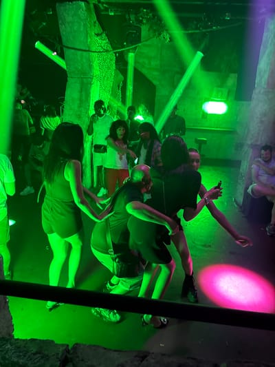 The image depicts a dimly lit indoor venue, likely a nightclub or event space in Canet-en-Roussillon, France, illuminated by vibrant green and pink lighting. A group of people are present, engaged in various activities. In the foreground, a person wearing a dark top and shorts is dancing, with their hands outstretched. To their left, another person in a dark top is also dancing. Behind them, a man in a black t-shirt and shorts is standing, and a woman in a white tank top and red pants is visible to his right, also dancing. Further back, more individuals are scattered throughout the space. The environment appears to be somewhat rustic, with rough-textured walls and what looks like a large, rough-hewn pillar or structure in the background, draped with netting. The overall atmosphere suggests a lively social gathering or party.