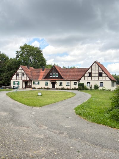 The image shows a large, two-story building with a traditional German half-timbered architectural style, featuring a red tile roof and white stucco walls with dark wooden beams. The building is set in a rural environment, with a large, well-maintained lawn in front. A gravel driveway splits and curves around the lawn, leading to the building. Trees and greenery surround the property, with a dense forest visible in the background. The sky is overcast with dramatic clouds. There are no people visible in the image. The building appears to be a residence or possibly a farmstead. The location is identified as Heiligenstadt i. OFr., Germany.
