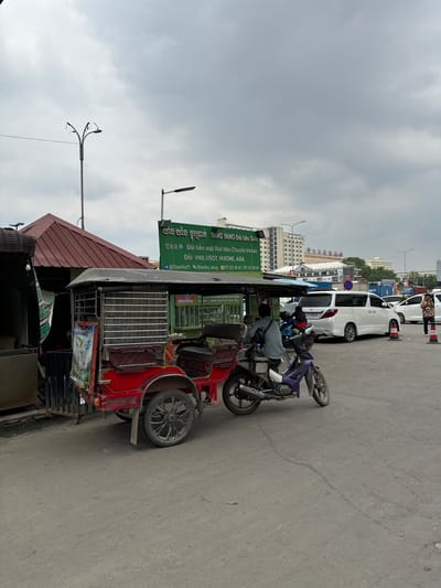The image shows a street scene in Bavet Quarter, Cambodia. A red tuk-tuk with a sign reading "YANG YANG" in multiple languages sits parked on the side of the street. The sign mentions money exchange services, including VND, USDT, and HUIONE, and lists phone numbers and a social media handle. A man wearing a gray shirt is partially visible on the tuk-tuk, possibly the driver. Behind the tuk-tuk, a purple motorcycle and several white vans are parked. Buildings and overcast skies are visible in the background. Street lights with multiple arms are visible in the foreground. The pavement is gray concrete.