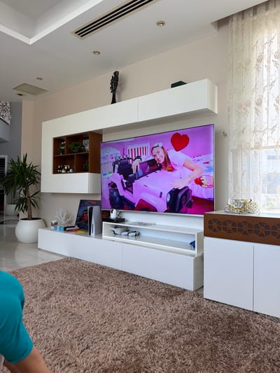 The image shows a modern living room with a large television displaying a video of a young woman in a pink outfit. The room is decorated with minimalist white furniture, including a media console and wall-mounted cabinets. A large brown shaggy rug covers the floor. In the background, a large window with sheer curtains is visible. A potted plant stands in the corner of the room. A person wearing a turquoise top is partially visible in the foreground. The scene suggests a comfortable and contemporary living space.