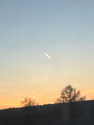 The image shows a landscape at sunset. The sky transitions from blue to orange/yellow near the horizon. There's a white, oblong object in the sky. At the right edge of the frame, a dark mass, possibly a tree line or hillside, is visible. The image appears to be taken from Novoromashkovo, Russia, because this is what the prompt says, but without independent confirmation.
