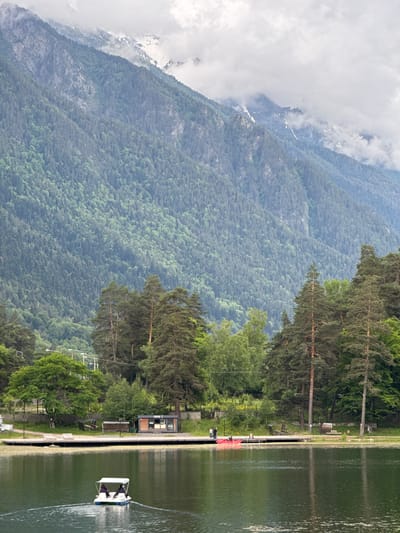 A wide shot captures a serene lake with a small pedal boat carrying two people in the foreground, creating ripples as it moves across the water. The lake is bordered by a wooden dock. On the dock, a few people are visible, along with a red kayak. Behind the dock, a small building with the word "COFFEE" is present, alongside benches and some trees. Further in the background, a dense forest covers the lower slopes of majestic, cloud-shrouded mountains. The scene is located in Verkhnyaya Teberda, Russia, as indicated by the surrounding natural landscape typical of the region.