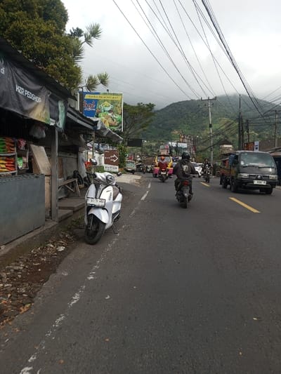The image depicts an outdoor street scene under an overcast sky in a mountainous region of West Java, Indonesia. This location is evidenced by visible vehicle license plates displaying the letter "F" (e.g., F 5315 WSI on a scooter, F 9390 YI on a truck), which corresponds to the Bogor region in West Java, Indonesia, and a brown directional sign pointing to "Telaga Warna," a known landmark in Puncak, West Java. The requested location of Saing Ta Mawt Lay, Myanmar, is not supported by the visual evidence.
The foreground features a paved asphalt road with a broken white line on the left and a solid yellow line on the right, indicating traffic flow. On the left shoulder, a white and brown Yamaha Grand Filano scooter (license plate F 5315 WSI, expiry 05-29) is parked facing right on its kickstand. Adjacent to the scooter, a single-story commercial structure with a corrugated metal roof and a black banner displaying "HZR PEDSHOP" is visible. Shelves outside this structure hold various packaged goods. Above the shop, a large billboard advertises food with the word "Bal," and a brown directional sign with a right-pointing arrow indicates "EXIT" and "Telaga Warna."
On the road, traffic consists primarily of motorcycles and a small truck, moving away from the viewer. In the mid-ground, a person wearing a black helmet and black hoodie rides a dark-colored scooter. Further ahead, multiple other motorcycles are visible, including one with a red body. A black Suzuki Carry pickup truck (license plate F 9390 YI) is present on the right side of the road. A yellow microbus is visible in the distance on the left. The background is dominated by a large, densely forested mountain under a cloudy sky. Numerous black electrical utility wires crisscross the upper portion of the frame, supported by visible utility poles.