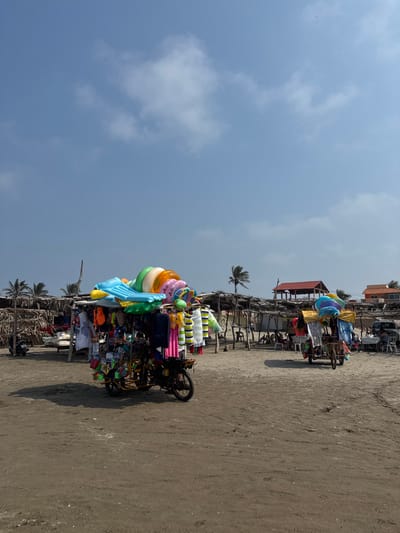 The image depicts a sunny outdoor scene on a sandy/dirt beach in Antón Lizardo, Mexico. The sky above is largely clear blue with scattered white cumulus clouds.
In the immediate foreground-left, a motorized three-wheeled vendor cart is parked, heavily laden with merchandise. The cart, appearing to be a modified motorcycle or tuk-tuk, has a dark frame and a lighter colored cargo section. On its roof, multiple inflated beach toys are stacked, including a light blue rectangular raft, several colorful donut-shaped swimming rings, and a green inflatable ring. Below, numerous colorful garments, likely swimwear or beach towels, are hung on display, along with smaller items such as plastic toys, sunglasses, and other accessories. A distinct shadow from this vendor cart is cast on the sandy ground to its right.
In the midground, a series of rustic, open-air structures with thatched roofs and wooden pole supports are visible, serving as shelters or market stalls. Under these shelters, several individuals are seated or standing, some at tables, indicating commercial or leisure activity. To the right of the foreground vendor cart, another similar, though less detailed, three-wheeled vendor cart is parked, also displaying inflatable items and merchandise.
Further in the background, a two-story building with a distinctive red tile roof and an open upper level is visible. To its right, another building with an orange-colored roof is partially obscured. Several palm trees are scattered throughout the midground and background, particularly on the left and center-right, characteristic of a coastal environment. The ground consists of uneven sand or packed dirt, with tire tracks visible, suggesting vehicular movement in the area. No direct body of water is visible within the frame.