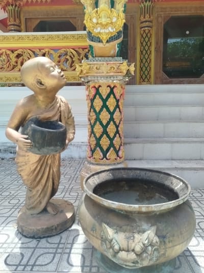 A bronze statue of a young monk stands on a tiled patio, holding a begging bowl. Next to it is a large, ornate bronze pot. Behind them, a temple entrance features white steps, gold and green decorative pillars, and wooden doors with glass panels. The sunlight suggests it is daytime. The scene appears to be in Thailand, given the temple architecture.