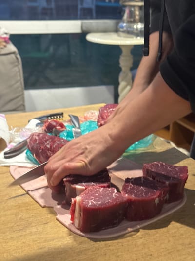 The image depicts a close-up view of raw beef steaks being prepared on a wooden surface. A person, whose arms are visible, is in the process of slicing one of the steaks with a knife. The steaks are thick-cut and appear to be tenderloin. Several other steaks are arranged on a light pink cutting board to the right. Various kitchen utensils, including knives and a corkscrew, are scattered around the preparation area, along with some crumpled plastic packaging. The background is blurred, showing a hint of a window with a view of an indoor setting with furniture, possibly a dining room or kitchen. The overall scene suggests food preparation activities taking place indoors. There is no indication of the specific location in Ptichnoye, Russia.