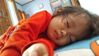 A young child, wearing an orange shirt, is sleeping on a bed with a blue and orange patterned bedspread. The child’s head is turned to the right, and the image is taken from the left side of the child. The child's eyes are closed, and their mouth is slightly open. The child's hair is dark and long. The child's right hand is visible, lying on the bed. There is a doorway to the left of the child, and a partially visible doorway to the right of the child. The room appears to be an interior space of a house. The coordinates provided indicate the location of the image is in the city of Kecamatan Panjalu, Indonesia.