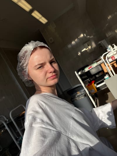 The image shows a young woman wearing a white disposable medical cap and a white garment, possibly a hospital gown or robe. She is seated in what appears to be a medical or clinical setting, with fluorescent lighting visible on the ceiling and medical equipment in the background, including shelves with various items, a trash receptacle, and what might be a medical cart. Sunlight streams in from the left, casting strong shadows across the scene. The woman is looking directly at the camera with a neutral expression. The environment suggests a sterile or treatment-oriented location.