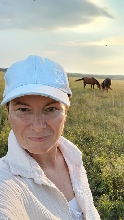 A woman with short, dark hair and light skin is shown in a close-up selfie taken outdoors. She is wearing a light blue baseball cap with a white logo and a white textured shirt over a white tank top. Her gaze is directed at the camera, and she has a slight smile. In the background, a field of green grass with scattered yellow wildflowers stretches to the horizon, under a pale, overcast sky. Two dark-colored horses are grazing in the distance. The setting appears to be a rural area, likely in Nadezhdenka, Russia, given the natural landscape and the presence of livestock. The lighting suggests it is either early morning or late afternoon.