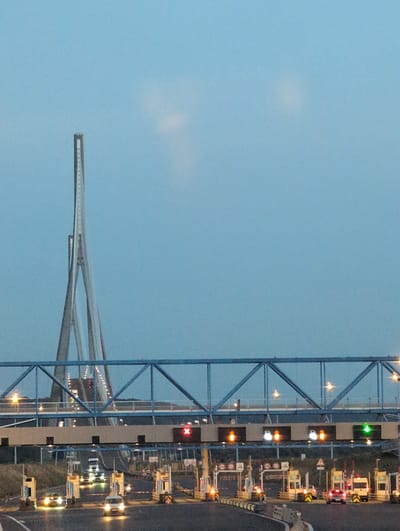 The image depicts a nighttime view of a toll plaza in Sandouville, France, likely at the approach to the Pont de Normandie. In the foreground, several lanes of traffic are visible, with cars and trucks illuminated by their headlights. Various toll booths are lined up, some displaying traffic signals. Above the toll plaza, a large blue metal structure, part of the bridge's infrastructure, spans across the lanes, dotted with lights. In the background, the distinctive, tall, A-shaped pylons of the Pont de Normandie rise dramatically against the pale blue, twilight sky. The overall scene captures a functional infrastructure element against a striking architectural landmark.