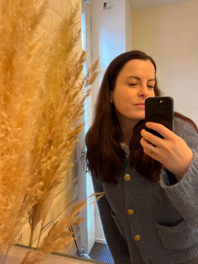 An adult female, reflected in a mirror, captures a self-portrait using a black smartphone held in her right hand. She has long, dark brown hair, fair complexion, and wears a light blue/grey textured knit cardigan with gold-colored circular buttons and two front pockets over a white garment. A two-band silver-colored ring adorns her left ring finger. The left foreground of the reflection is dominated by an out-of-focus arrangement of dried, yellowish-brown pampas grass. The background reveals a bright, minimalist interior with light-colored walls, possibly white rectangular tiles. A tall, narrow window or glass-paneled door frame is visible, admitting light with a blue hue, and a dark, striped floor mat lies near a threshold. A small glass diffuser bottle is reflected on a surface beneath the pampas grass. A ceiling-mounted device, resembling a security camera, and a minor wall imperfection are visible in the upper reflected background. This interior scene is located within Kaiserslautern, Germany, consistent with a modern commercial space such as a cafe or boutique.