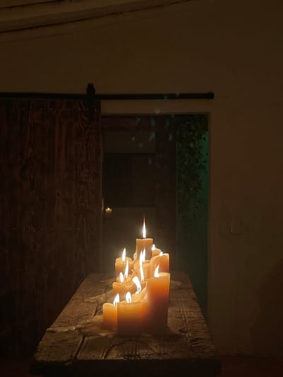 A lit candelabra rests on a wooden beam, with flames casting light on the scene. The wooden beam is textured with visible grain, and the wall behind is dimly lit. Through the open door, green foliage is visible, suggesting an outdoor space. A darker wooden structure and what appears to be another candle sit near the foliage. The door has a simple latch mechanism.