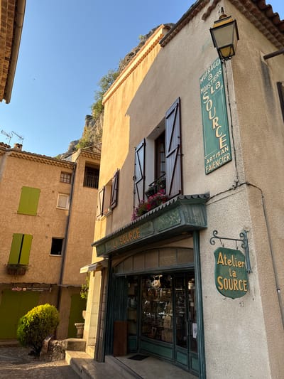 The image depicts a narrow cobblestone street in the village of Moustiers-Sainte-Marie, France, bathed in the warm light of late afternoon. The buildings are constructed of pale, textured plaster and feature terracotta tiled roofs. The focal point is a shop with a green-framed glass storefront, displaying various items inside. Above the shop entrance, a green sign reads "LA SOURCE." A vertical green sign on the building's corner, adorned with a streetlamp, displays "MUFRAGGI A MOUSTIERS La SOURCE ARTISAN FAIENCIER." Another hanging green sign reads "Atelier la SOURCE." Shutters on the buildings are a mix of dark wood and bright green. A small, leafy green bush grows from a stone planter on the left. The background reveals a rugged, rocky cliff face. The sky is a clear, vibrant blue.