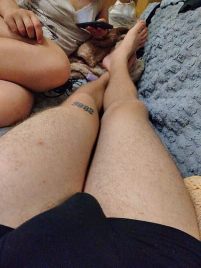 The image is a close-up, low-angle shot of a person's legs and torso, taken from a seated position. The subject is wearing dark-colored shorts and is resting on a fuzzy, grey blanket. To the left of the frame, another person's legs are visible, wearing a white t-shirt with text and graphics. One hand of this second person is visible, holding a dark smartphone. A small, light-brown dog or stuffed animal is partially visible between the legs. A tattoo of the number "2002" is visible on the left thigh of the main subject. The scene appears to be indoors, possibly a living room. There is no information in the image that can be used to determine the location in Okhtyrka, Ukraine.