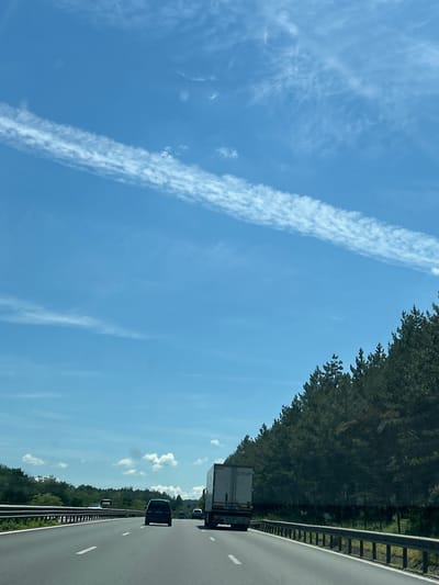 This image depicts a highway scene, likely on the road towards Novi han, Bulgaria, during daylight. The sky is a clear blue with wispy white clouds. On the right side of the highway, a dense forest of pine trees lines the road. Several vehicles are visible on the highway, including a dark-colored SUV, a semi-trailer truck, and other cars in the distance. The road is asphalt with clear lane markings. The environment suggests a rural or semi-rural setting outside of a major urban area. There are no people clearly visible in the image.