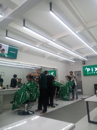 The image displays the interior of a modern barbershop named 'PIX BARBER SHOP' in Sindanglaya, Indonesia. The brightly lit space features a white ceiling with a grid of exposed beams and several black-framed, suspended LED strip lights. The walls are light-colored. A prominent feature is a large mirror extending across the left side, above white counters.
Five individuals are visible:
1. **Barber 1:** Standing in the left-center, a male with bleached blonde hair, wearing a dark green polo shirt and black trousers, is actively cutting the hair of a client using clippers.
2. **Client 1:** Seated in a black barber chair, facing away, this young male is draped in a green patterned cape featuring the words "BARBER SHOP" and a repeating monogram.
3. **Barber 2:** In the right-center, a male wearing a white and black striped polo shirt, dark trousers, and a black face mask, is cutting the hair of a second client using scissors.
4. **Client 2:** Also seated in a black barber chair, facing away, this young male is draped in a green patterned cape identical to Client 1's.
5. **Barber 3:** A third barber-client interaction is visible as a reflection in the mirror, indicating another male barber in a dark shirt attending to a client.
On the upper left wall, a digital screen displays the word "MULLET" alongside an image of a person with a mullet hairstyle. A large, illuminated green sign with white text, partially visible on the right wall, identifies the establishment as "'PIX BARBER SHOP". The floor is covered in white hexagonal tiles with a contrasting dark gray strip running through the center. A white table with a black frame is present in the lower right foreground.