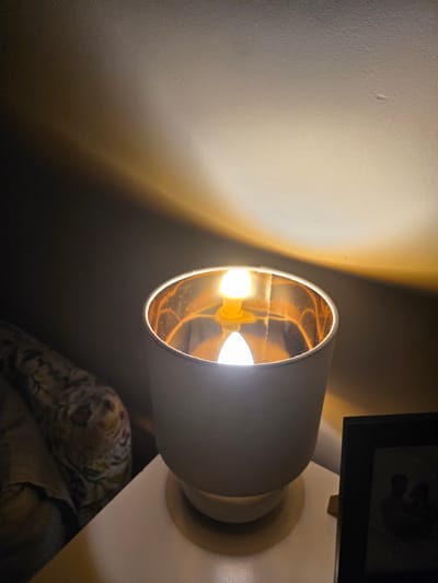The image shows a close-up view of a lamp on a nightstand. The lamp has a white exterior and a reflective gold interior. A lightbulb is visible inside the lamp, emitting a warm glow. The light casts a prominent shadow on the wall behind the lamp. To the left of the lamp, a portion of a floral-patterned pillow or blanket is visible. To the right of the lamp, a black picture frame is partially visible, containing a dark silhouette of what appears to be two people. The scene suggests an indoor setting, likely a bedroom, and is illuminated by the lamp. The image does not provide any specific information or context related to the city of Brzeg, Poland.