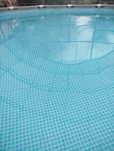 The image shows a close-up view of an above-ground swimming pool filled with clear blue water. The pool liner has a pattern of small, light blue squares. Reflections of what appears to be a structure, possibly a greenhouse or patio roof, are visible on the water's surface. In the upper left corner of the original image, a glimpse of people and part of the pool's frame can be seen. The framing appears to be a grey metal structure with white connectors. The scene suggests a sunny day, given the reflections and the brightness of the water. The setting is likely a residential area or recreational space in Krupietski Selsovet, Belarus. There is no visible text in the image.