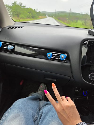The image provides a first-person perspective from inside a car, looking forward through a wet windshield. The scene depicts travel through a rural area, potentially Бабстово, Russia, characterized by lush green, hilly landscape, a visible road, and power lines under an overcast or rainy sky.
In the foreground, a person's left hand is prominent, with a simple gold ring on the ring finger. The fingernails feature bright alternating colors: pink on the index and little fingers, and orange on the middle finger. The hand is casually posed with the index and middle fingers extended upwards, possibly a peace sign. A cheetah-print wristband is visible around the wrist. Below the hand, a leg is seen, dressed in light blue jeans and a grey sneaker, resting in the car's footwell.
The car's dark dashboard and central console are visible, featuring air vents. Three triangular blue car air fresheners, each emblazoned with the white text "AMMY," are clipped into the air vents. The central console includes various buttons and a dial labeled "TEMP." On the right, the car's side pillar displays the text "SRS AIRBAG." The wet windshield suggests ongoing or recent rain, contributing to the soft, natural light in the vehicle.