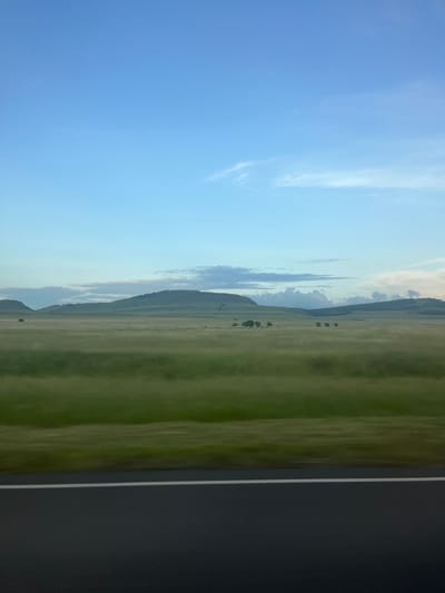This is a landscape photograph taken in Dzhirim, Russia. The image features a vast, green, open field with subtle striations indicating freshly cut grass or tilled earth. In the distance, rolling hills rise, shrouded in a light mist or haze that softens their contours and creates a serene, atmospheric effect. The sky above is a clear, bright blue, transitioning to a lighter hue near the horizon, with delicate wisps of clouds scattered across its expanse. The overall impression is one of natural beauty and tranquility, likely captured during a clear day, possibly in the morning or late afternoon given the soft lighting. The absence of any people or man-made structures in the field emphasizes the untouched, natural state of the landscape. The far right edge of the image shows a dark, out-of-focus area that might be a wall or a shadow.