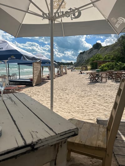 The image depicts a beach scene in Topola, Bulgaria. In the foreground, a weathered white wooden table and chair are visible, casting shadows on the sandy ground. Beyond this, a line of beach umbrellas, some featuring the Corona logo, shades the sandy beach. The sea, with its turquoise water, stretches out to the horizon, where a line of trees and a rocky cliff are visible. Several beach loungers and partitions made of woven reeds are set up along the sand. A few people are scattered on the beach, some seated and others walking. The sky is filled with dramatic white clouds against a clear blue expanse.