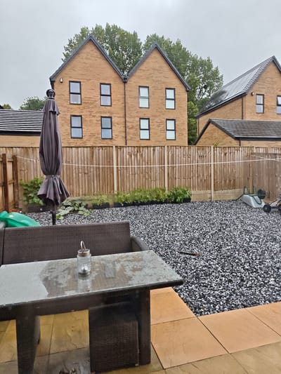 This is an outdoor shot of a backyard in the UK, possibly Locking Parklands, taken on an overcast day, as indicated by the muted light and slightly damp appearance of the table and patio stones. The scene depicts a residential backyard with a gravel ground cover interspersed with several small, potted plants lined up along a wooden fence. Behind the fence, two modern, multi-story brick houses with dark-tiled roofs are visible, along with a line of trees. In the foreground, a wicker sofa and a glass-topped coffee table with a small lantern on it are positioned on a patio area made of large, square, orange-toned paving stones. A folded patio umbrella stands nearby, to the left of the frame. Further into the yard, a green plastic slide for children and a grey wheelbarrow are present. The overall atmosphere is calm and appears to be after a rain shower, with water droplets visible on the table. There is no visible text in the image.