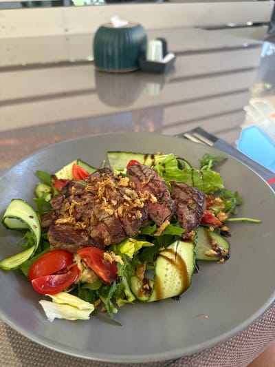 The image shows a close-up of a salad with steak on a gray plate. The salad contains sliced cucumbers, cherry tomatoes, and leafy greens. The steak is sliced and topped with crispy fried onions. A dark sauce, likely balsamic glaze, is drizzled over the salad and steak. The plate is on a table with a woven placemat. In the background, slightly out of focus, is a teal-colored tissue holder and a salt and pepper shaker. The setting appears to be an outdoor restaurant or cafe. The image was taken in Shirokaya Schel, Russia.