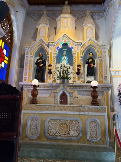 The image depicts an altar within a church in Hội Tín, Vietnam. The altar is a multi-tiered structure, predominantly yellow with white ornamental carvings. At the center of the altar, a statue of the Virgin Mary, wearing a blue robe, is positioned under a canopy adorned with colorful lights. Flanking the Virgin Mary are two statues of male figures in religious attire. To the left, a figure in a black robe stands holding a ladder. To the right, a figure in a black robe holds a red book.

Below the central statues, two wooden lamps with white spherical globes on ornate bases are placed. The lower section of the altar features decorative carvings, including a circular emblem with initials that appear to be "IM". The surrounding walls are painted in muted colors and show signs of age. To the left of the altar, a stained-glass window with red, yellow, and blue panes is visible. In the foreground to the left, a dark wooden furniture piece, possibly a pew or confessional, is partially in view. The floor is covered with patterned tiles. A red velvet rope barrier is present on the right side of the image. The overall scene suggests a religious setting with traditional architectural and decorative elements.