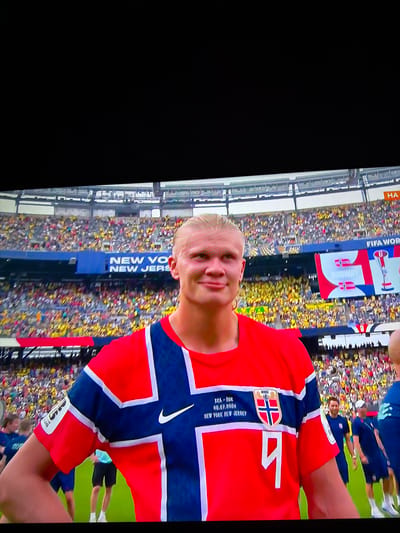 A male athlete, likely a soccer player, stands on a green field in a stadium, wearing a red and blue Norwegian national team jersey with the number '9' on it. He has blonde hair pulled back and is smiling at the camera. Behind him, a large crowd fills the stadium seating, which is predominantly yellow and blue. A large screen displays "FIFA WORLD CUP" and the Norwegian flag, along with trophy graphics. The text "NEW YORK, NEW JERSEY" is visible on a blue banner above the crowd. The lighting suggests it is daytime. The location is a stadium.