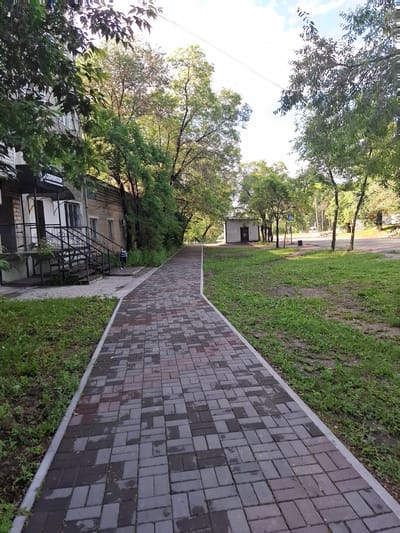 The image shows a paved walkway in Svobodny, Russia. The walkway is constructed with interlocking rectangular pavers in shades of grey and reddish-brown. To the left of the walkway is a brick building with a dark entrance and metal stairs. Green grass and trees are visible on both sides of the walkway. In the distance, a small white building with a dark door can be seen, along with a pedestrian crossing sign. The sky is partly cloudy. The scene depicts an outdoor urban environment.