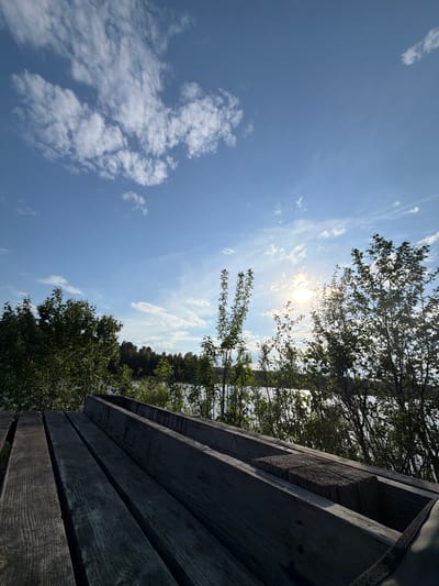 The image depicts a view towards the sky from what appears to be a wooden structure. The sky is predominantly blue with scattered white clouds. The sun is visible, shining through a canopy of trees. Part of the wooden structure is in the right portion of the frame, showing horizontal planks with visible wood grain. Some trees are visible in the background. The image is oriented with the wooden structure on the right and the sky and clouds on the left. The location is in Selychka, Russia.