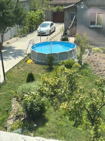 The image shows a residential yard in Чистенское сельское поселение, Ukraine, on a sunny day. The yard contains a large, above-ground swimming pool filled with blue water and a silver ladder. Beside the pool on the grass, there is a yellow inflatable object and some other items. A white car is parked on a concrete driveway in the background, next to a grey house with a window. The yard is surrounded by green grass, trees, and shrubs. In the foreground, a tree with green leaves and what appear to be small fruits is visible. Several solar-powered garden lights are placed in the grass. The overall scene depicts a typical suburban or rural outdoor setting.