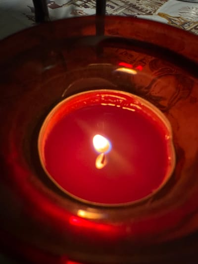 Profesor Ishirkovo, Bulgaria: A close-up, overhead view depicts a single red candle burning actively within a dark red, cylindrical glass candle holder. The candle's red wax is partially melted, encasing a visible wick that produces a bright, white-yellow flame with a faint blue base. The flame illuminates the immediate surroundings with a warm, reddish glow. The inner surface of the reflective red glass holder displays several distinct reflections: a faint, distorted profile of a human head or face is visible on the right side, along with two distinct, parallel horizontal red light streaks on the upper left. Subtle reflections of a light-colored patterned textile, consistent with a tablecloth, are also visible. The background, outside the immediate vicinity of the candle holder, is out of focus, showing sections of a patterned textile surface and dark, vertical elements, possibly furniture legs. No direct human presence or interaction is observed beyond the reflection.