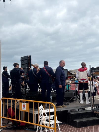 Captured from a slightly elevated perspective, the image shows a musical performance in the city of Arrecife, Spain. The band consists of approximately six musicians dressed in dark clothing, playing on a stage. A man in a traditional white and red outfit stands behind them, on the second row of the stage. One man is dressed in a blue shirt and trousers and is standing at the front of the stage. A crowd of people is visible in the background, watching the performance. The sky overhead is overcast. In the foreground, a yellow metal barrier separates the stage from the viewing area. The city's emblem or logo is displayed on the barrier. A variety of equipment is on stage, including speakers, microphones, and instruments.