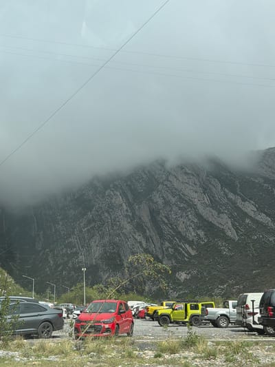 The image captures a mountainous landscape partially shrouded in fog, with a line of vehicles parked on the right side of the frame. A large, rugged mountain or cliff face, textured with rock and sparse vegetation, dominates the background, with a thick layer of fog or cloud obscuring its upper and central portions.
In the foreground, a row of cars is parked facing to the left, perpendicular to the main image orientation (which is rotated). Prominently visible are a red car, followed by a bright yellow-green off-road vehicle (likely a Jeep), a white pick-up truck or SUV, a dark grey SUV, and a black SUV at the end of the line. These vehicles appear to be parked on a paved or gravel roadside. To the right of the parked cars, there's a strip of grassy and rocky ground with small bushes. A few light poles or street lamps are visible behind the parked cars. Overhead, multiple thin lines, possibly power lines or cables, stretch across the pale, overcast sky on the left side of the image.
The weather appears foggy or cloudy, especially impacting the visibility of the mountain. The lighting suggests it is daytime, but overcast. There is no discernible human activity or visible text in the image. The scene is set in Santa Catarina, Mexico, featuring a natural and developed roadside environment.