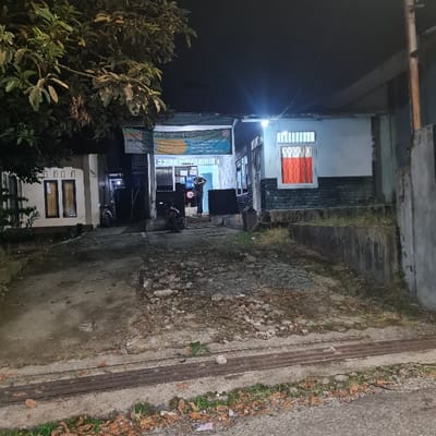 The image shows a nighttime view of a street in Kecamatan Tamansari, Indonesia, at coordinates -6.638 latitude and 106.735 longitude. The street is paved with a rough, uneven surface. A person is walking through an open doorway of a building on the left side of the image. Two motorcycles are parked on the left and right side of the driveway in front of the building. The building is lit by a streetlight overhead, and the walls are painted white.  A sign with lettering in the Indonesian language is displayed above the building.  There are trees to the left of the building, and a small fence and a grassy area to the right.  There is a small structure next to the building on the left, with a single window with bars on the side.  The building on the right side of the image has a closed window with an orange curtain.  The windows are lit from the inside of the building.  The ground is covered in dried leaves and debris.
