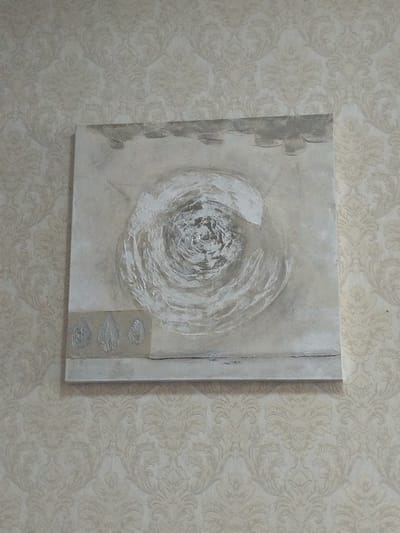 A square canvas painting with a textured, abstract design is mounted on a wall with ornate damask wallpaper. The painting's primary colors are shades of beige, gray, and white. The central motif is a swirling, circular pattern with a rough, impasto texture, reminiscent of a vortex or a stylized flower. The upper portion of the painting features a band of muted gray with irregular, cloud-like shapes. The lower portion has a horizontal band of light beige with three textured leaf-like imprints, arranged symmetrically. The overall style is minimalist and contemporary, with a focus on texture and subtle color variations. The wallpaper behind the painting has a repeating damask pattern in a slightly lighter shade of beige, creating a subtle contrast with the artwork. The lighting appears to be indoor ambient light, casting soft shadows. No people are visible in the image. The location context of Temse, Belgium, does not offer specific visual cues within the provided image.