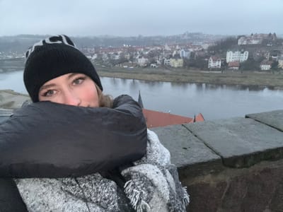 The image displays a person in the foreground observing a panoramic river and townscape under an overcast sky. The subject, a young individual, is visible from the chest up, looking directly at the viewer with a neutral expression. They are dressed for cold weather, wearing a black beanie with a white, indistinct pattern or text, a dark, possibly padded, jacket or coat, and a grey and white patterned scarf.
The immediate foreground features a grey stone or concrete ledge, indicating an elevated viewpoint. Beyond this, a wide river or body of water dominates the mid-ground, its surface reflecting the subdued light. The far bank of the river is lined with sparse vegetation.
Across the river lies a historic-looking town, identified as Meissen, Germany. Numerous buildings with varied architecture and distinctive red-tiled roofs are visible, sprawling across the landscape. A prominent structure with a dark red roof stands closer to the river. The sky is a uniform light grey, creating a hazy, muted atmosphere that suggests an overcast and likely cold day. No specific activity beyond observing the view is evident. The white pattern or text on the beanie is stylized and not clearly legible.